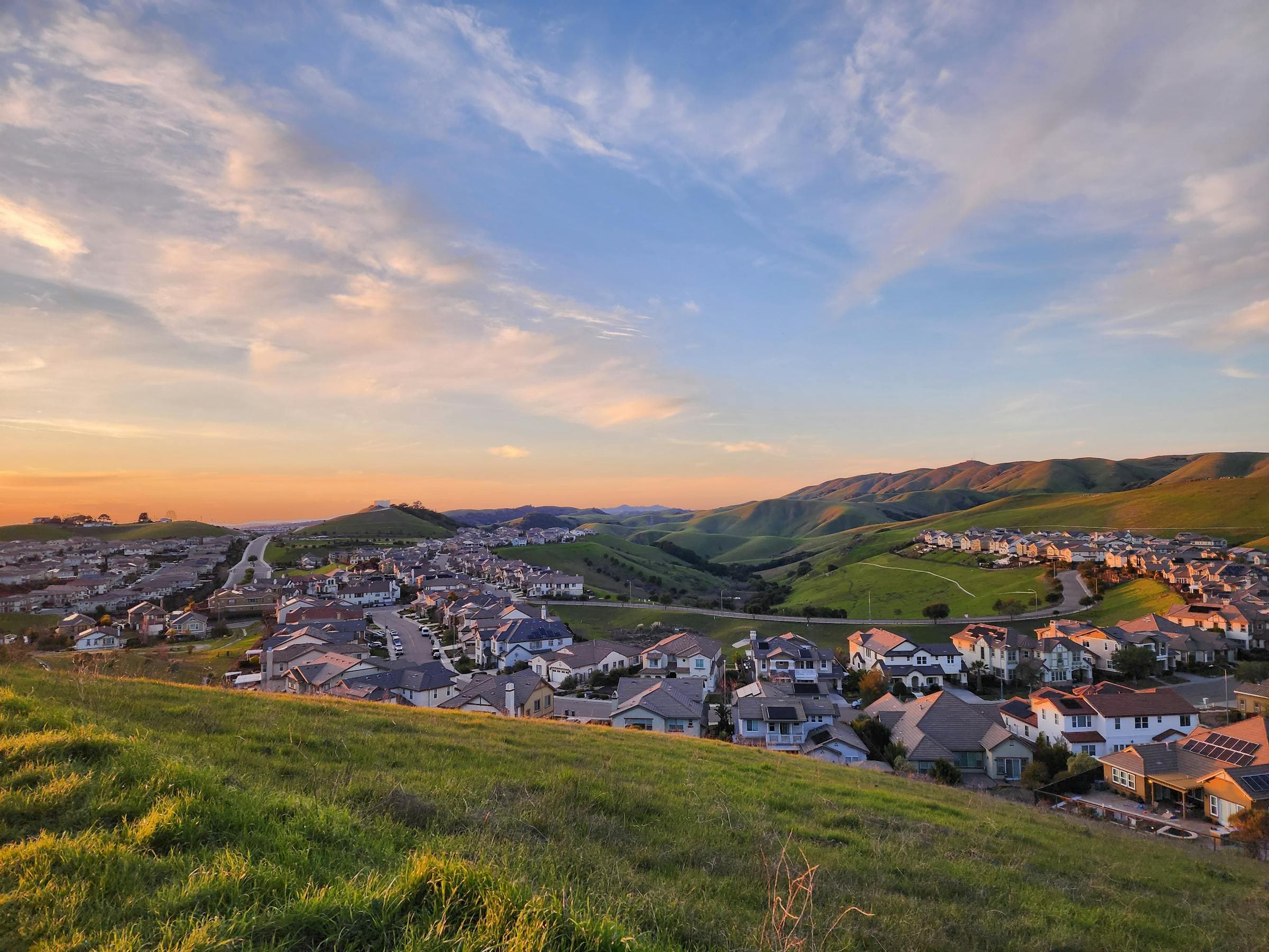 Hillside view of a Southern California suburban neighborhood at golden hour, with red-roofed homes nestled between rolling hills under a warm sunset sky