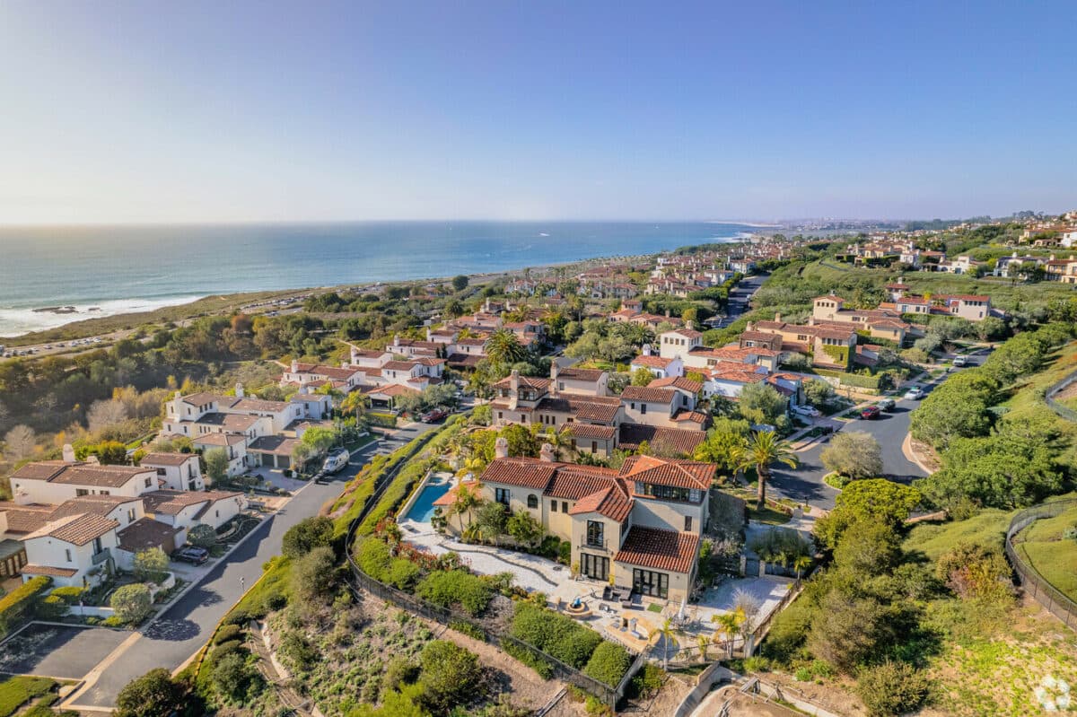 Aerial view of tile-roofed Newport Coast estates and a pool terrace on the hillside above Pacific Coast Highway, with the Crystal Cove shoreline and the Pacific beyond