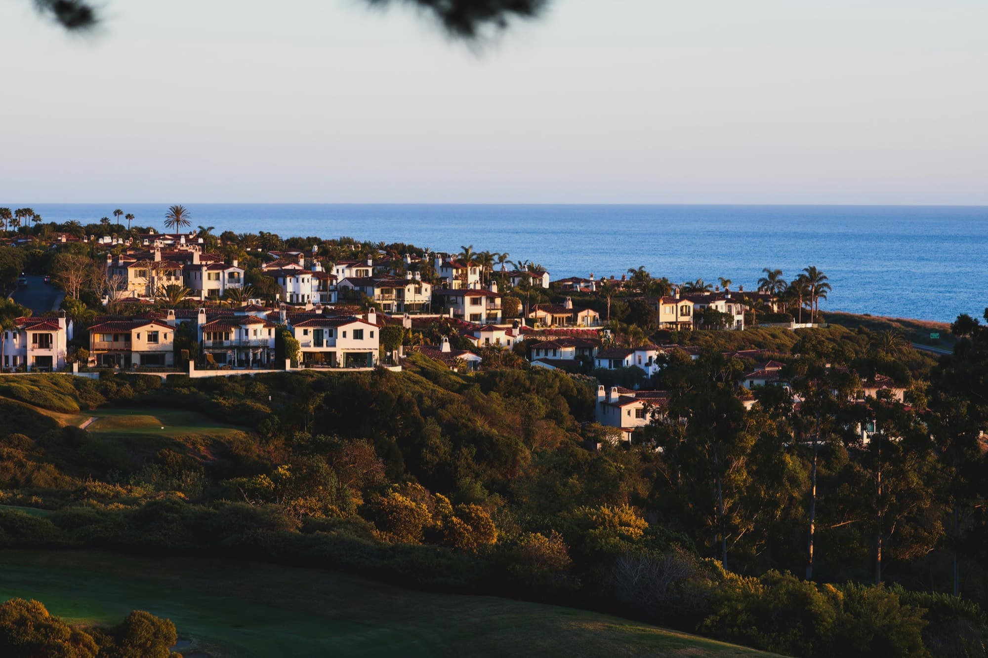 Mediterranean-style homes with red tile roofs lining a green hillside above a golf fairway, with the Pacific Ocean behind them at golden hour
