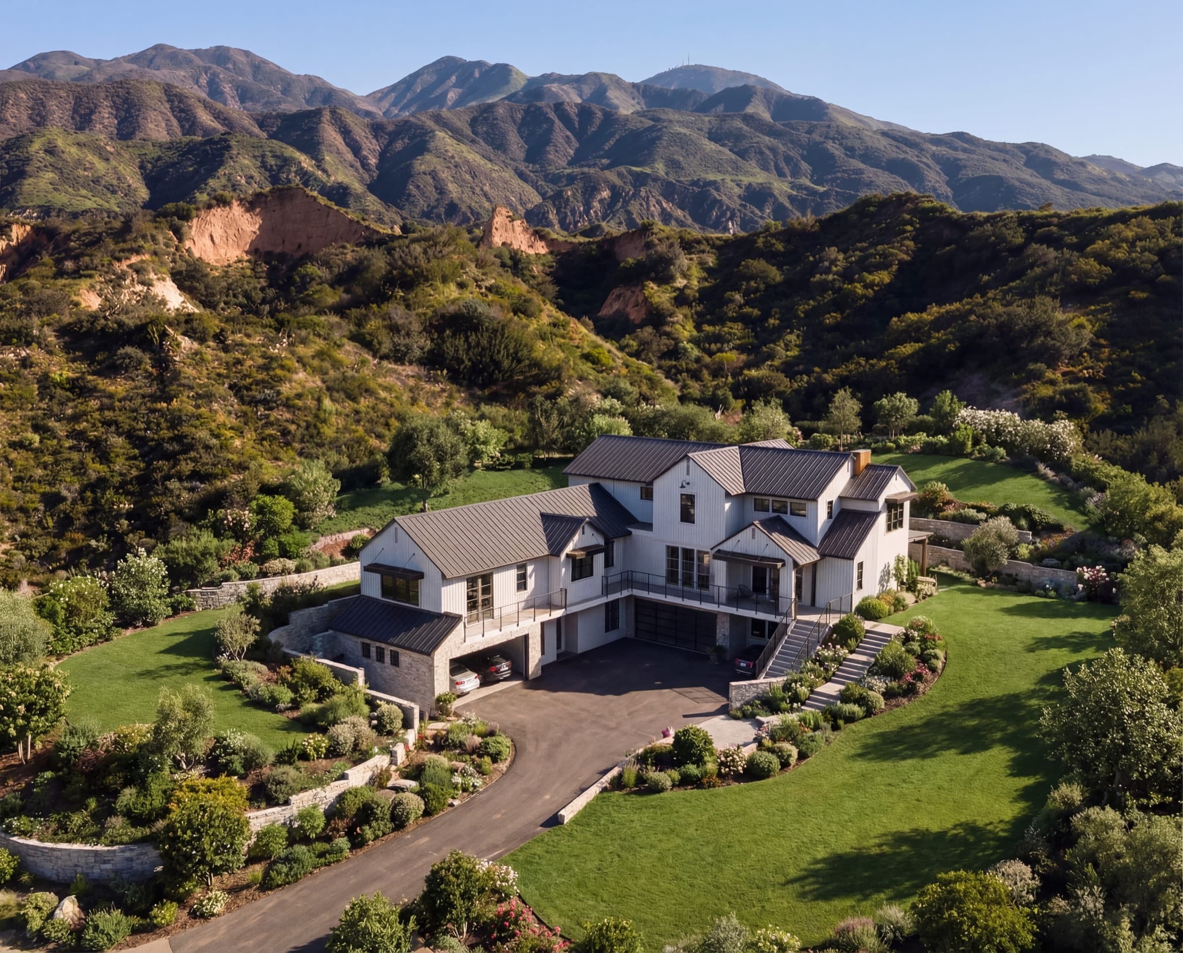 Aerial view of a white farmhouse-style home on a landscaped hillside lot with green foothills and mountains behind