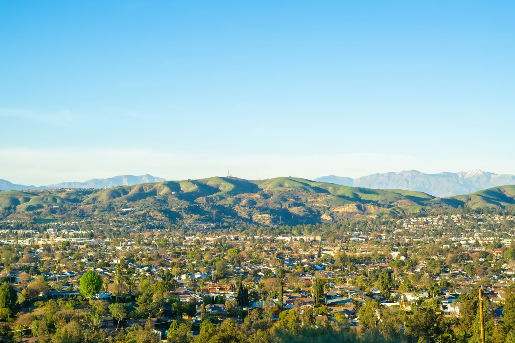 Orange County neighborhoods spreading across a valley below green hills, with snow-capped mountains on the horizon