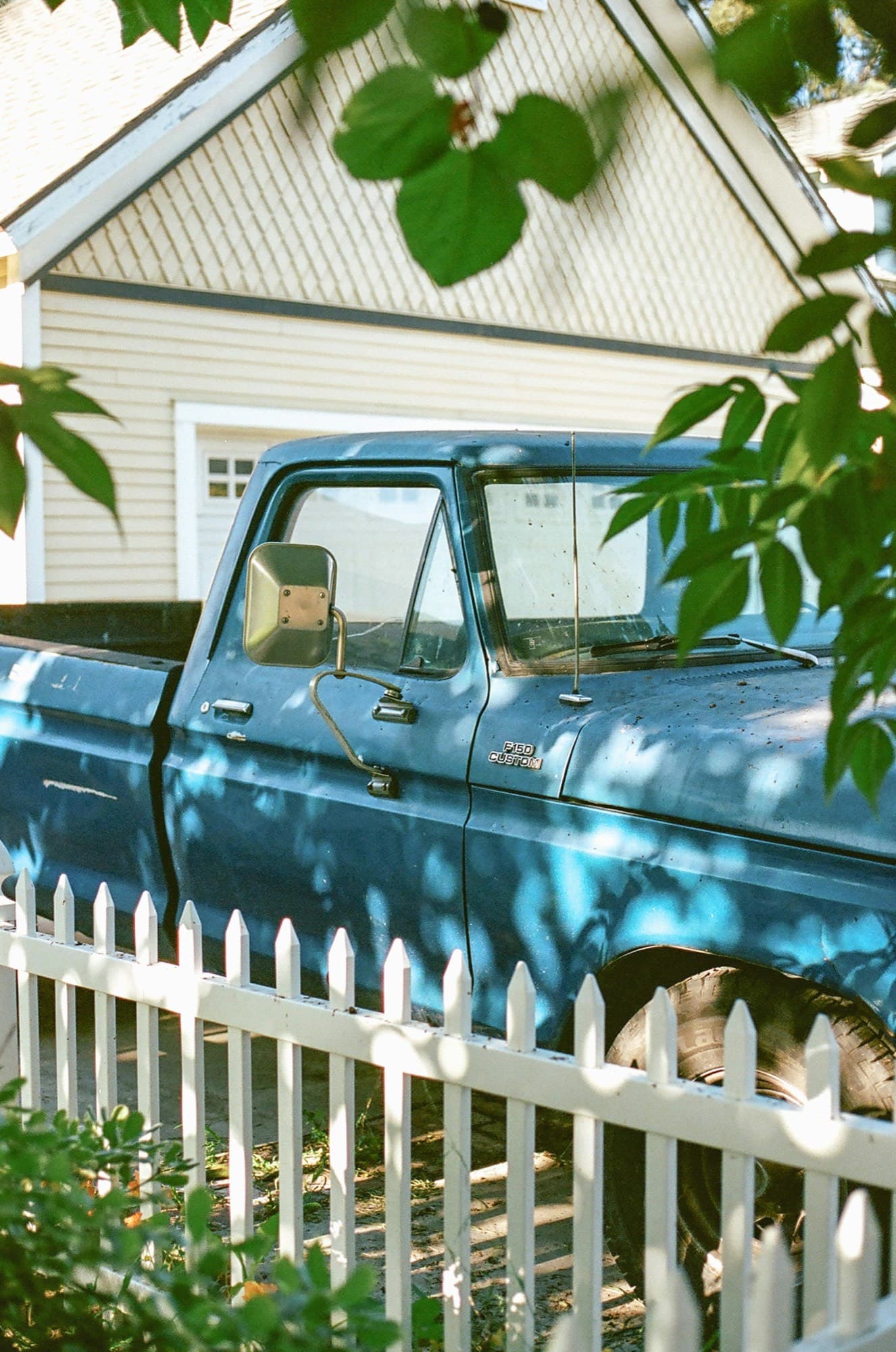 Vintage blue Ford pickup parked behind a white picket fence in front of a clapboard house in Tustin